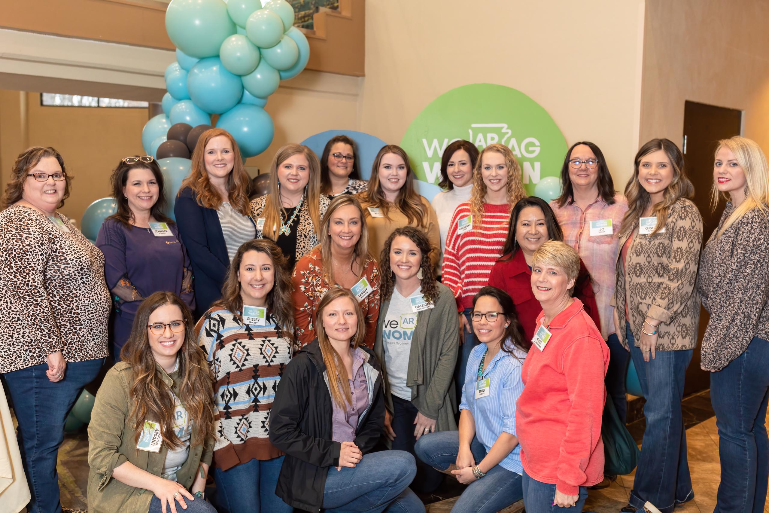 16 smiling ladies standing in front of teal balloons at the AR Women in Ag conference. 