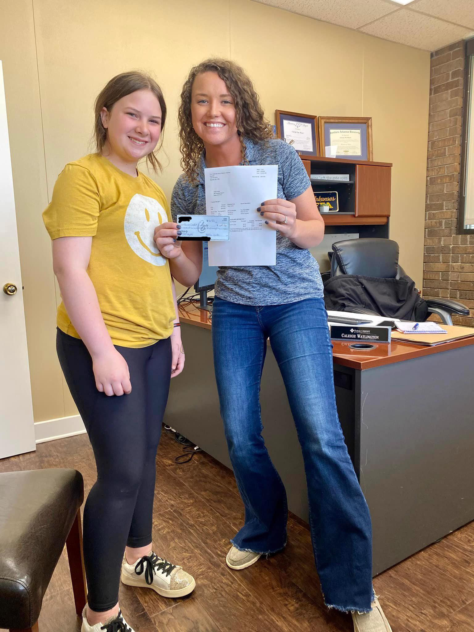 young girl in yellow shirt with happy  face on it smiles as Loan Officer Caleigh Watlington shows her paid off youth loan paperwork. 