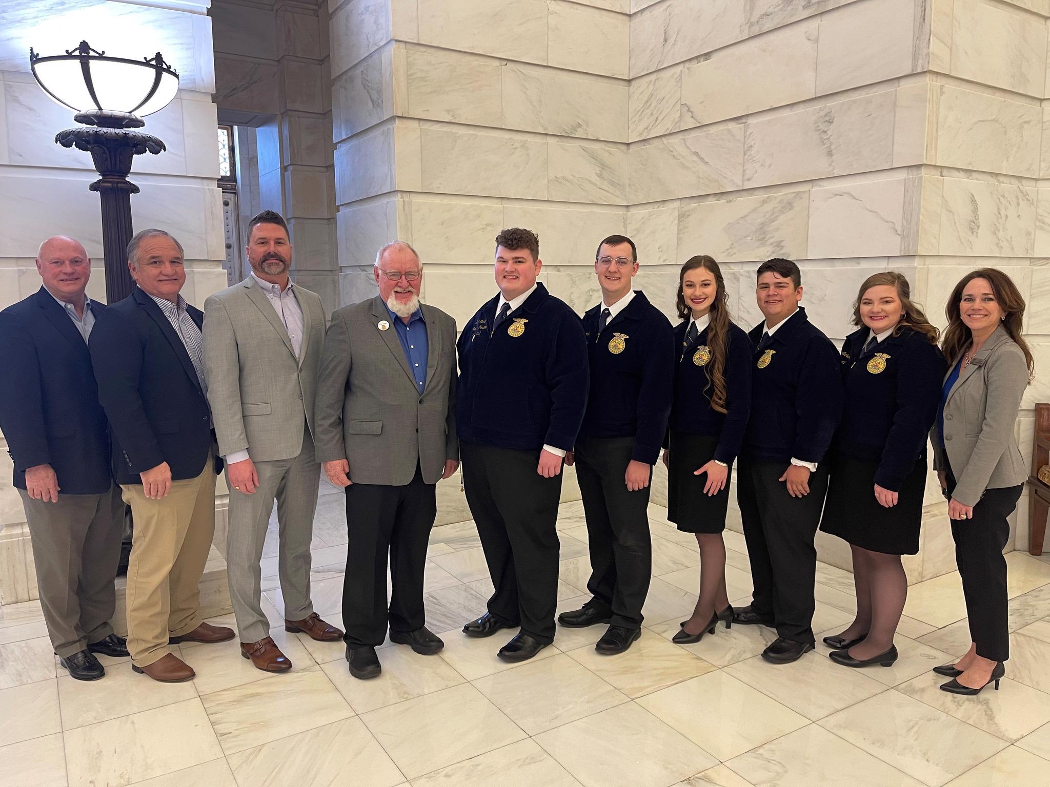 6 people standing in front of a white marble wall at the AR State Capitol.  Four on the right are wearing official FFA jackets and photo recognizes Farm Credit's $50,000 donation to updating Camp Couchdale. 